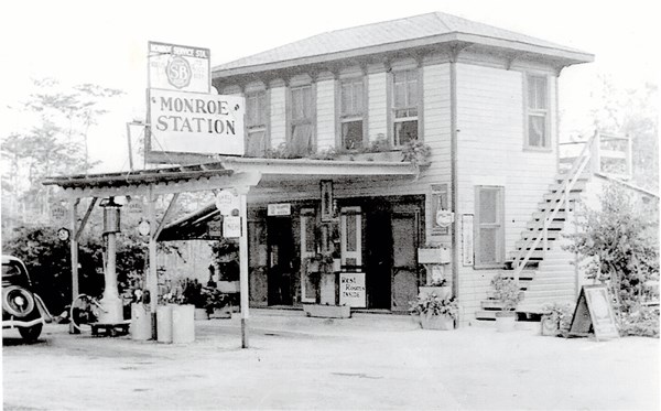 A two story store with gas pumps outside, and stairs on the right. A car is parked at the gas pumps and a sign above the pumps reads, "Monroe Station."
