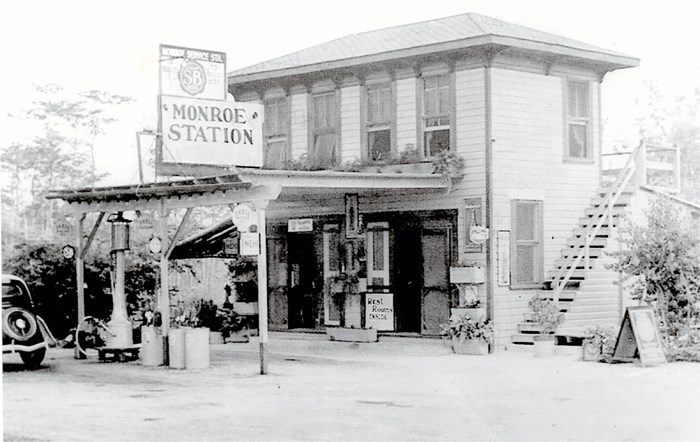 A two story store with gas pumps outside, and stairs on the right. A car is parked at the gas pumps and a sign above the pumps reads, "Monroe Station."