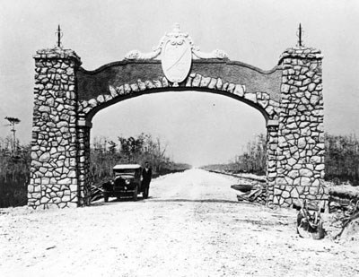 A man stands next to a car on a dirt road under a stone arch.