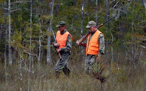 Two hunters in camouflage and blaze orange carry rifles.