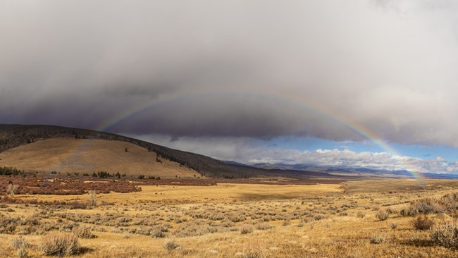 View of the Nez Perce encampment at Big Hole National Battlefield. Rain clouds loom above and a rainbow strikes across the image.