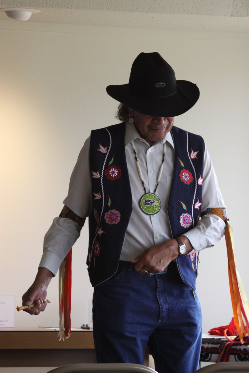 Leroy Seth shares his expertise as a Cultural Presenter in the Summer Speaker Series. Portrait of a man standing turned toward the camera. He is wearing a black cowboy hat, a white collared shirt, a beaded necklace, a black vest with a beaded floral pattern, and blue jeans. Ribbons are tied at his elbows and flow towards the ground.
