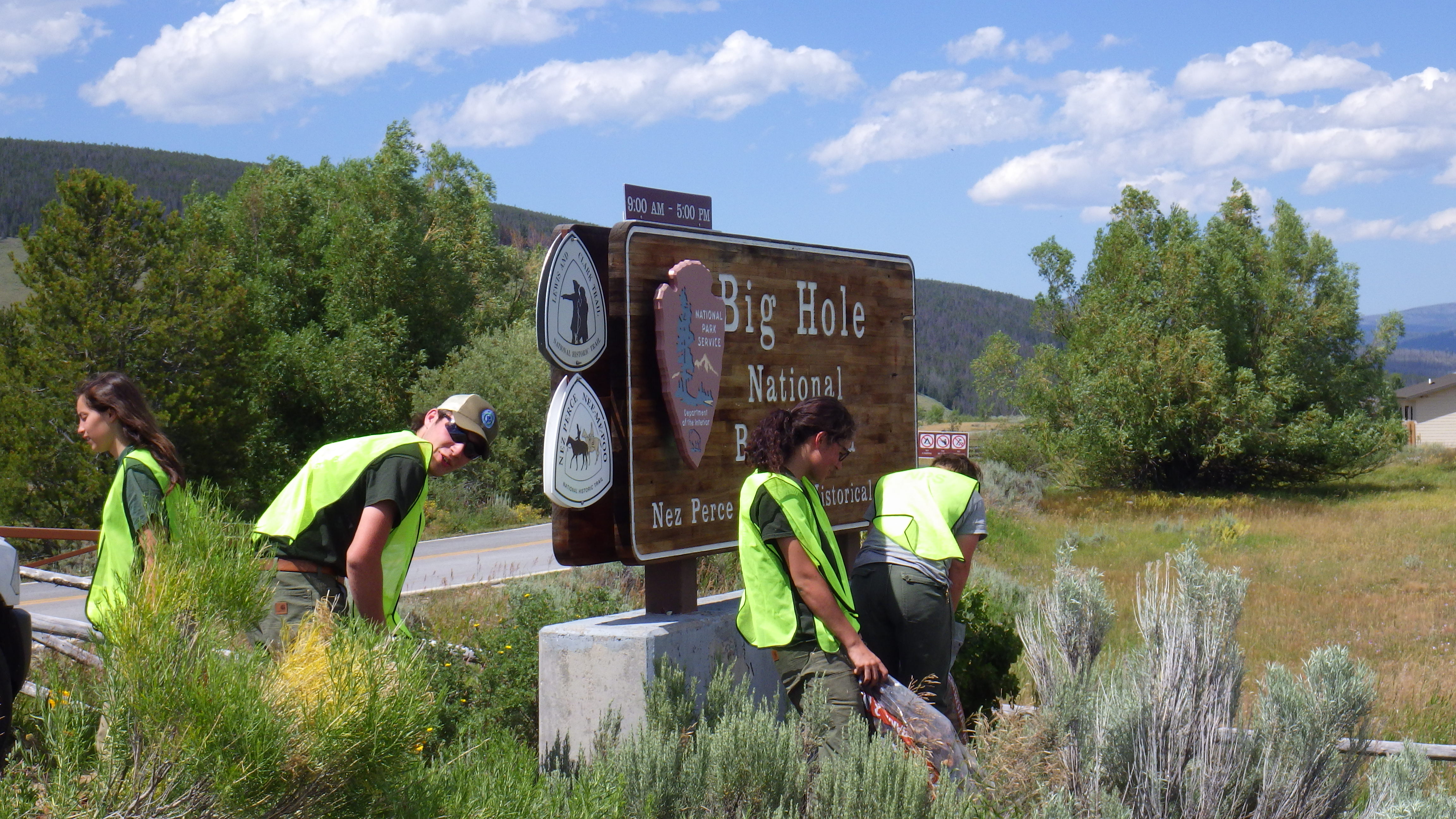 4 people wearing neon safety vests stand in tall grass around a sign reading Big Hole National Battlefield