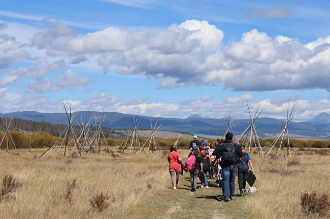 A park ranger leads a group of 15-20 children and adults on Big Hole National Battlefield's Nez Perce Camp Trail on a partly cloudy day. They are walking towards a group of tipi poles in a field of tall, dry grass with hills and mountains in the distance.