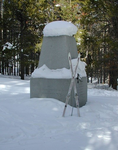 A granite monument in the snow with a pair of cross country skis next to it.