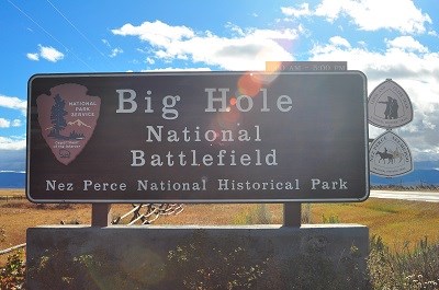 A wooden sign with the words "Big Hole National Battlefield Nez Perce National Historic Park" carved onto it with the the National Park Service, Nez Perce National Historic Trail, & Lewis & Clark National Historic Trail logos attached.