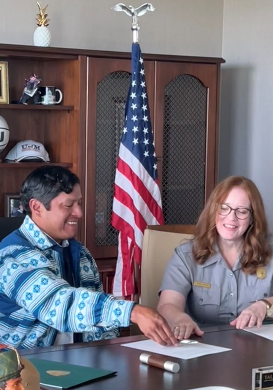 From left to right: A male tribal leader dressed in traditional Miccosukee jacket sits next to a uniformed park ranger. They are behind a desk in an office.