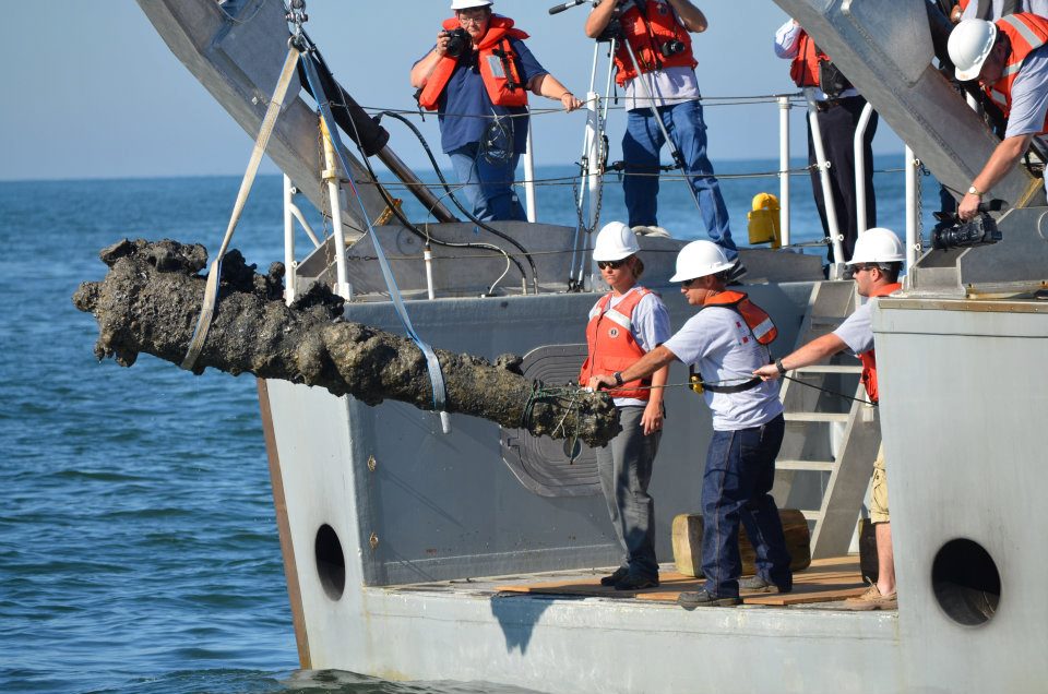 A historic cannon is raised by winch onto a boat by workmen.