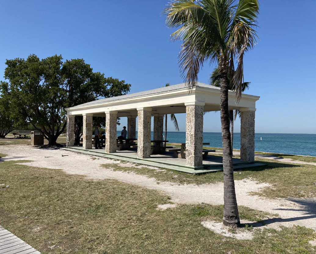 Boca Chita Pavilion Image shows a pavilion constructed out of white stone. Ten pillars and multiple picnic table. The image is flanked by grass, palm trees, blue water, and blue sky.