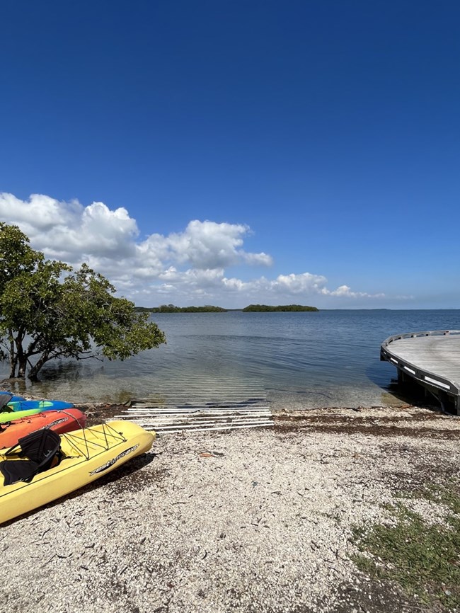 A canoe and kayak launch made of gravel and plastic tubes leads into calm blue water. Kayaks are seen on the left and a boardwalk on the right/