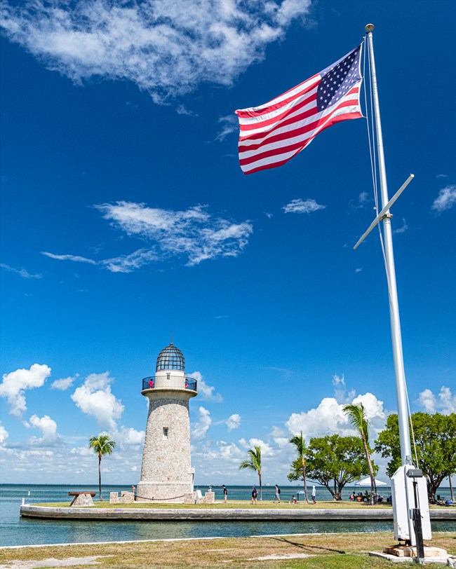 A high-flying American Flag dominates the right side of the image, standing in bright sunlight and a brisk wind. To the left is a picturesque lighhouse. People and palm trees populate the grounds. Beyond are a bright blue sky and calm turquiose waters