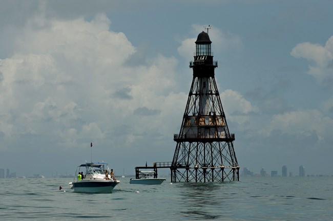 Weathered old lighthouse surrounded by bay waters with boats anchored around it and city skyline in the background