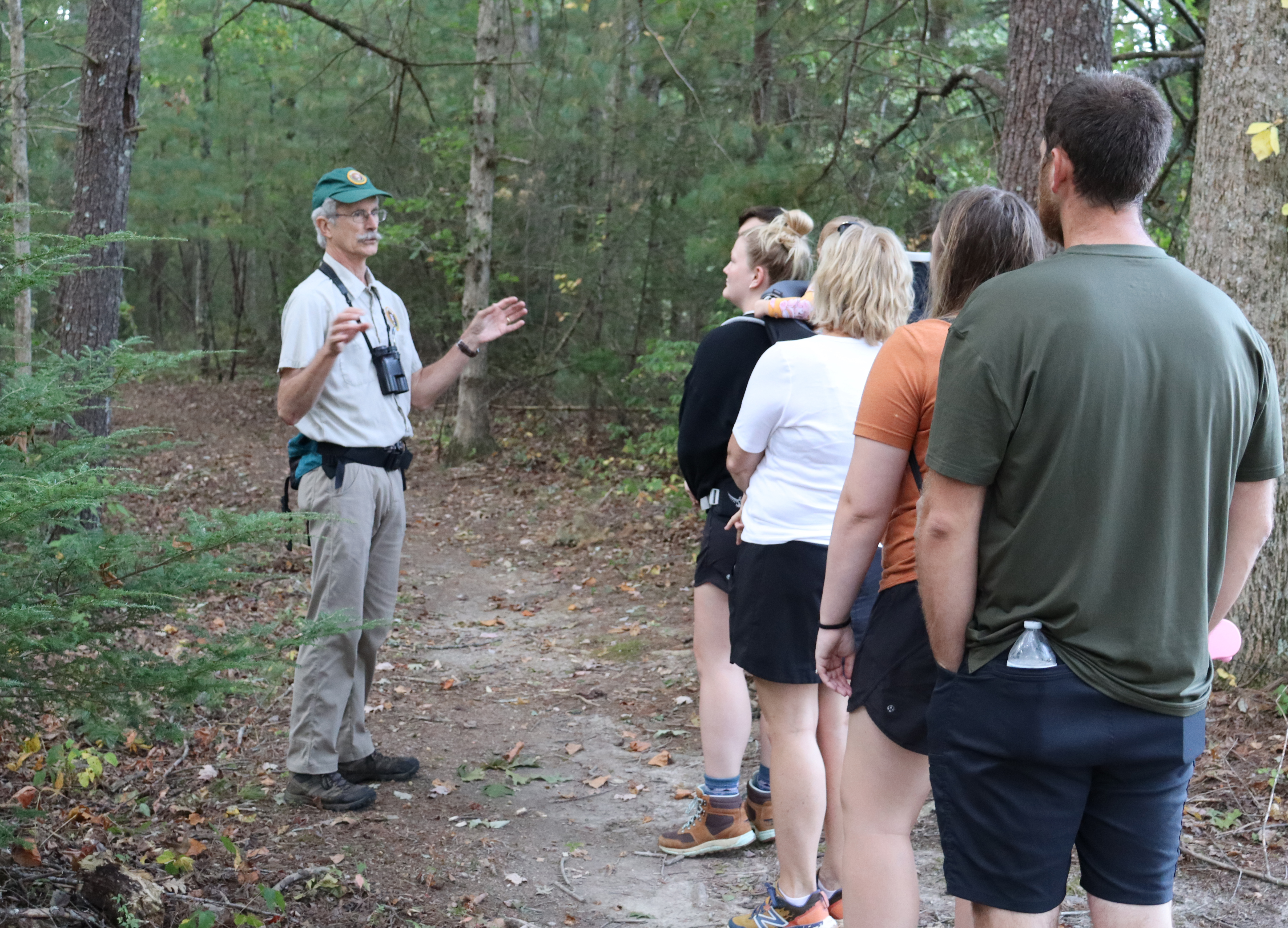 A man leads a small group of people on a guided bird hike on a trial in the woods.