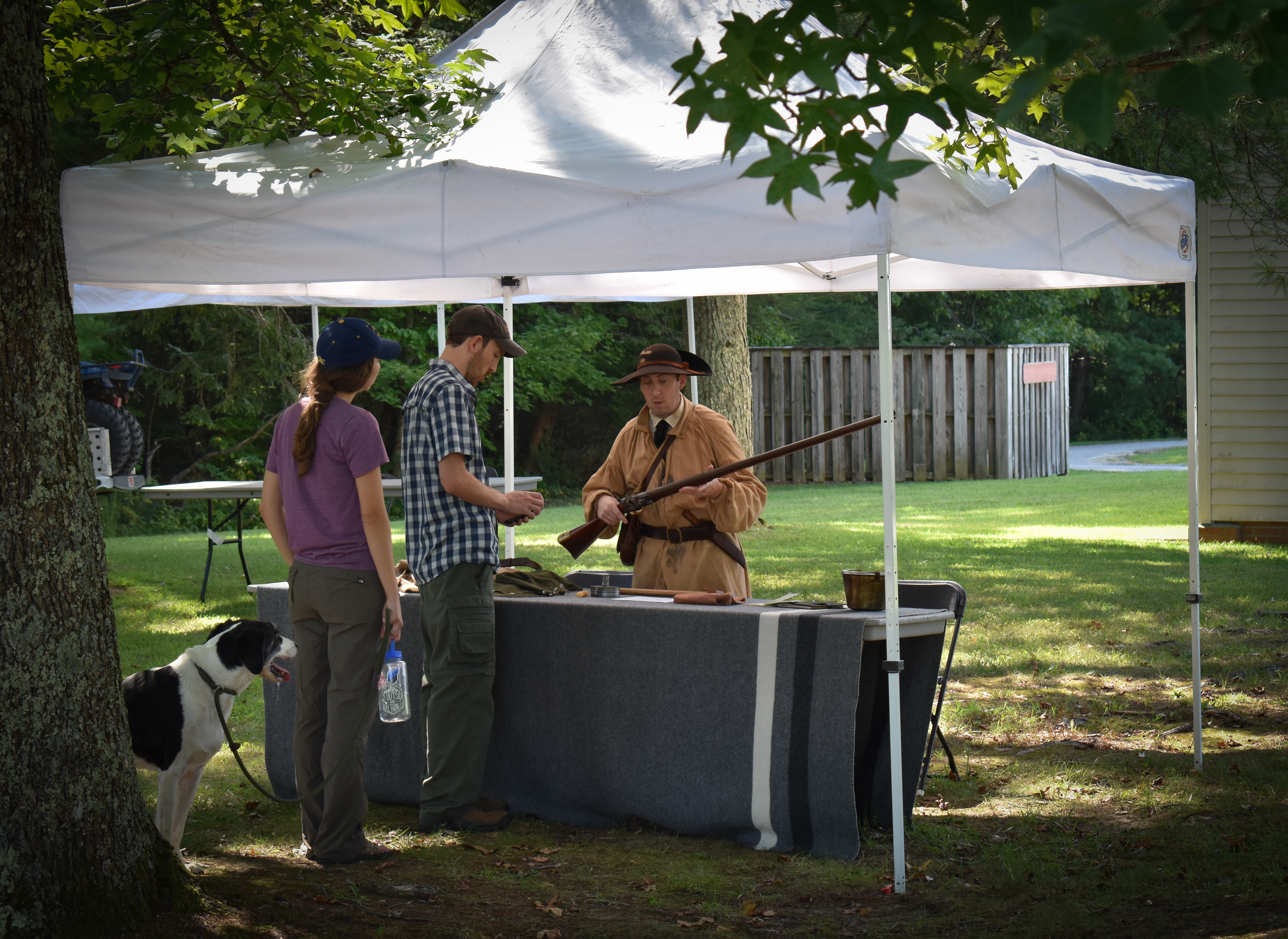 Long Hunter Demonstration A historical reenactor demonstrates musket use to two visitors under a white tent. A woman with a dog watches attentively. Outdoor setting with trees.