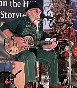 an older man sitting on stage playing a guitar and singing