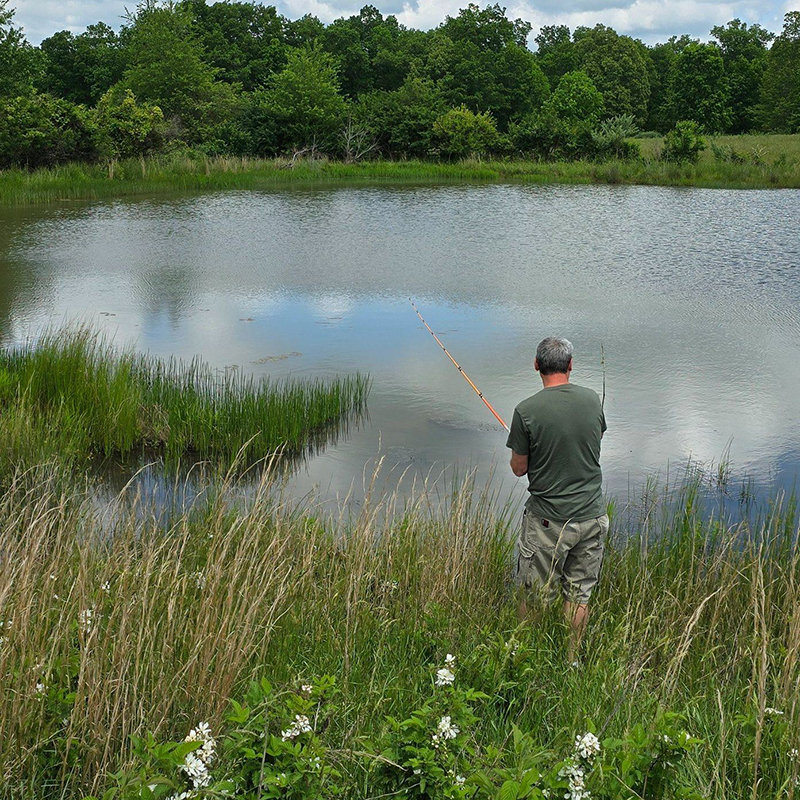 Fisherman standing in tall grass and wildflowers, fishing near water.