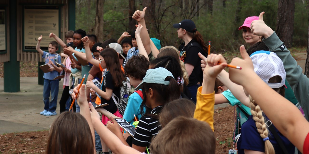 Students on a hiking adventure are giving a thumbs up. Many have pencils in their hands.