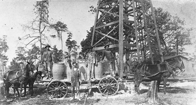 men standing next to mule drawn wagon beneath oil derrick