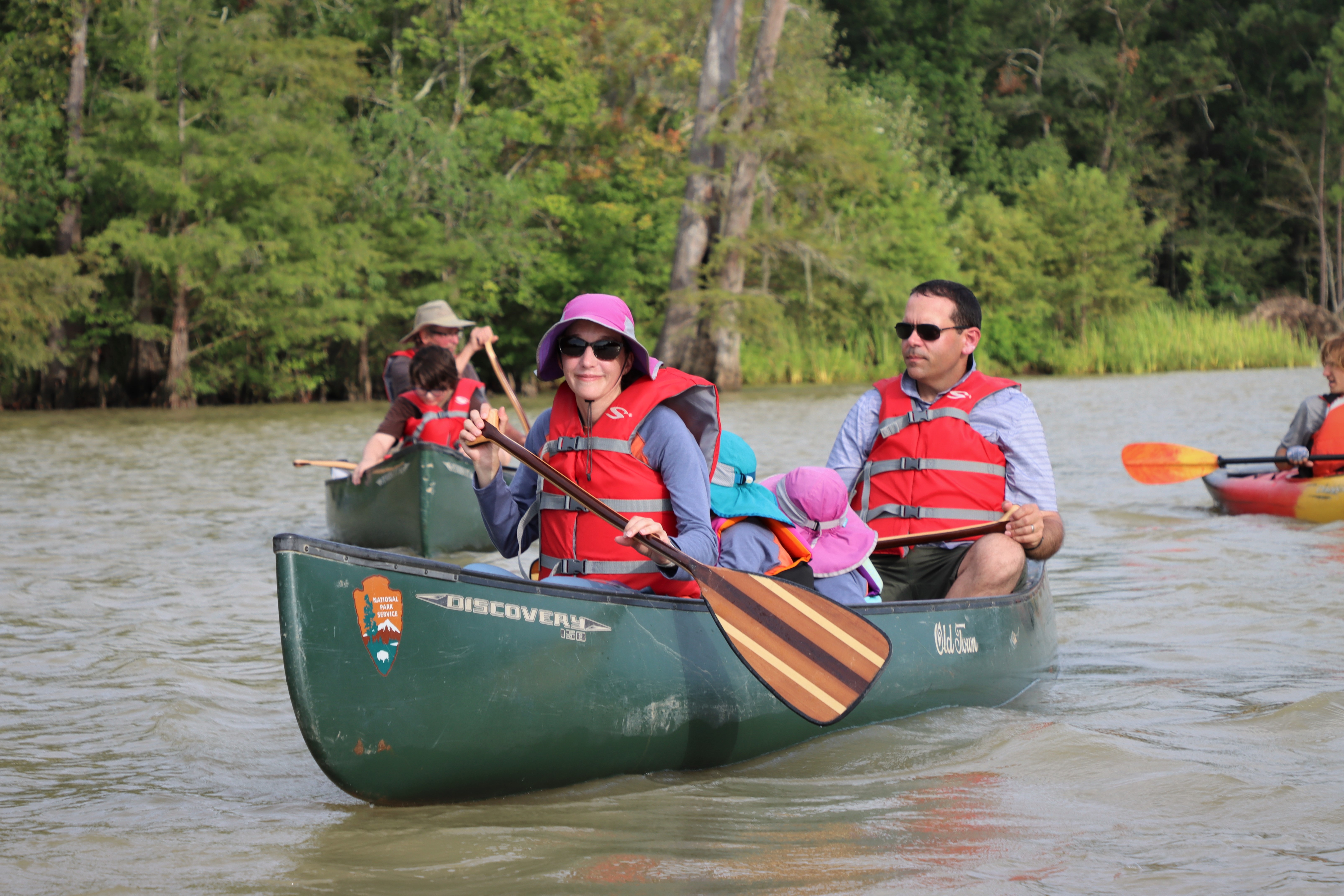 A family of 4 paddles a green canoe on a wide river. Each person is wearing a red life vest and holding a wooden paddle. There is another canoe in the background being paddled by 2 people and a kayak with 1 person inside.