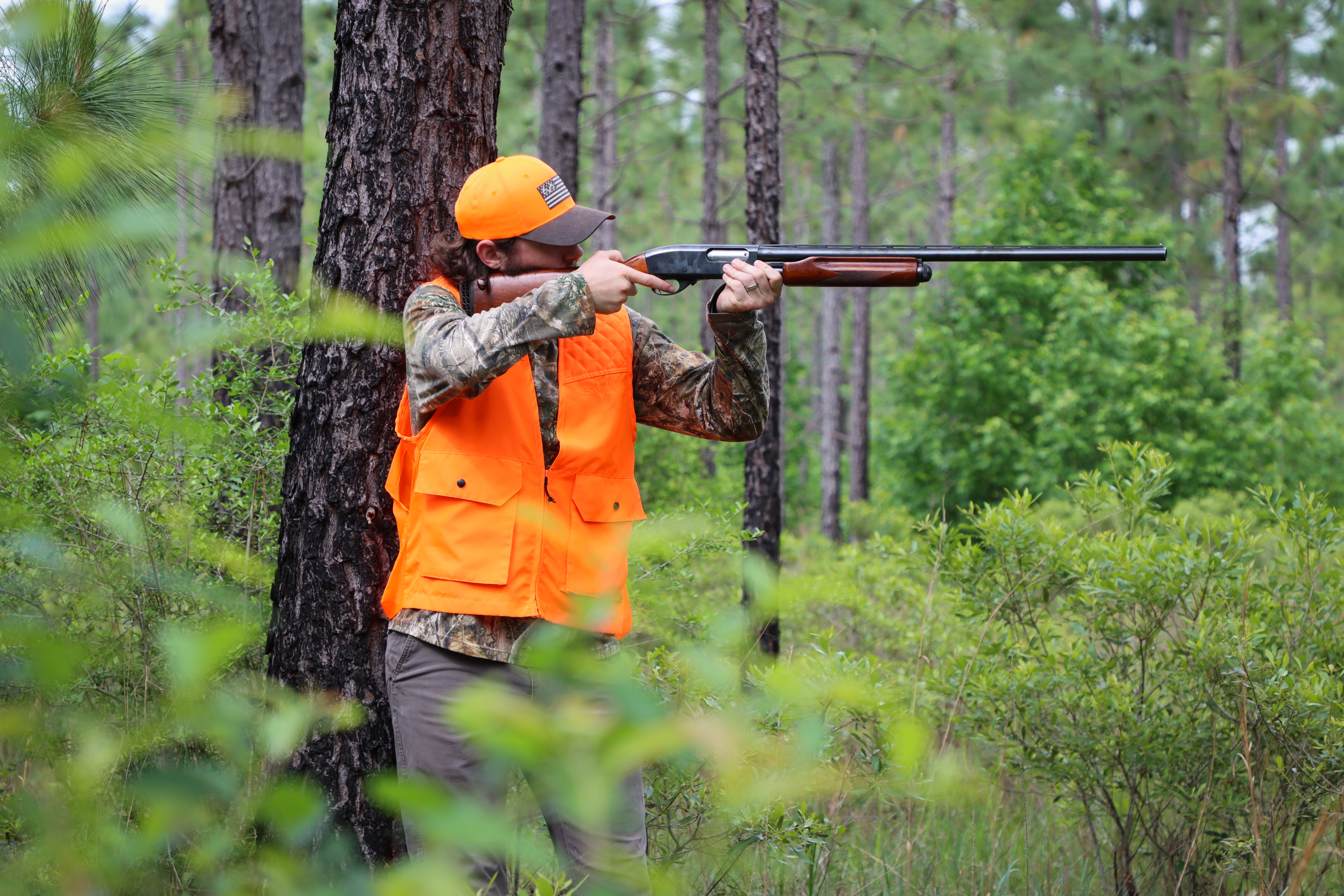 a male hunter wearing camo and orange vest and hat aiming a shotgun while standing in a green forest.