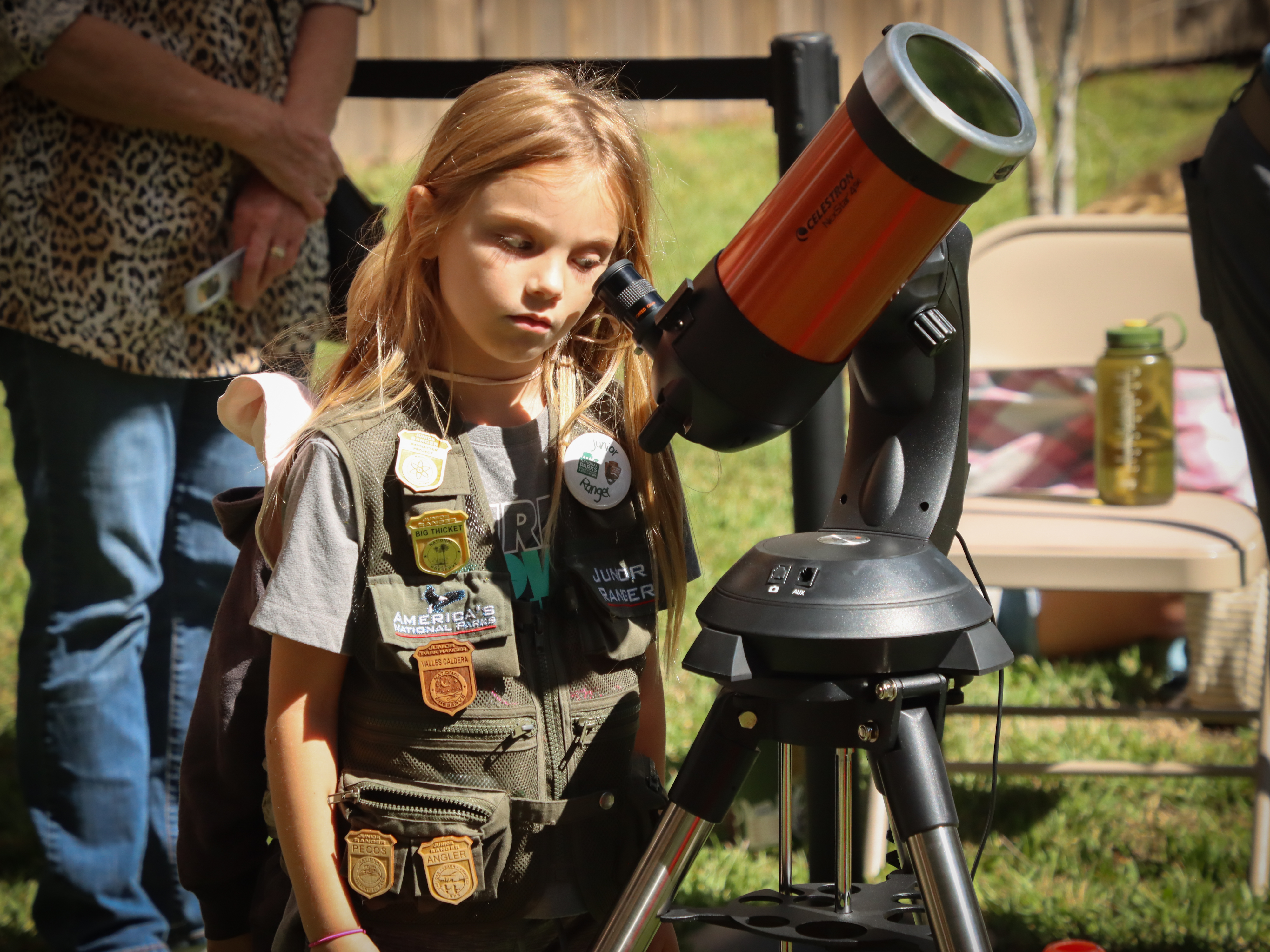 a young girl wearing a junior ranger vest decorated with junior ranger badges looks through a portable telescope equipped with a solar filter.