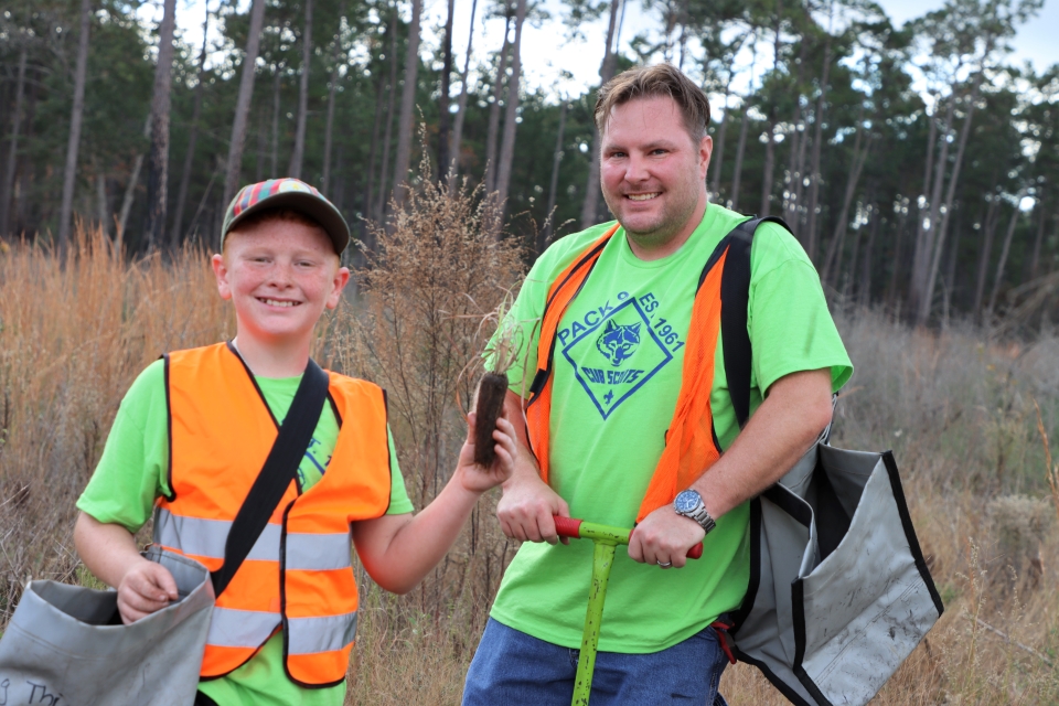 a father and son, wearing orange vests, hold up grass seedlings and planting equipment outside in the woods
