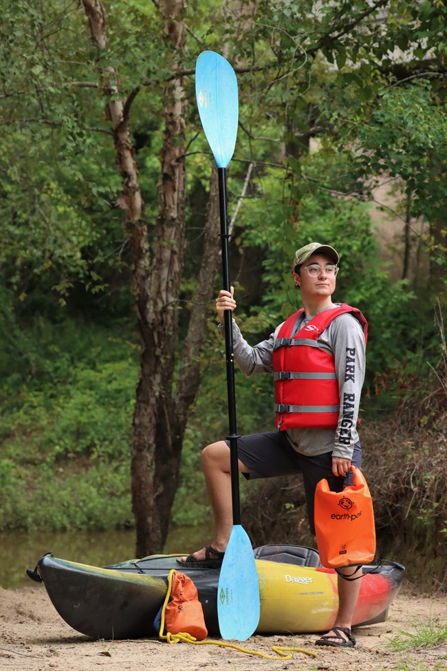 Paddle Intern CJ posing with paddle, life jacket, kayak, and other safety gear