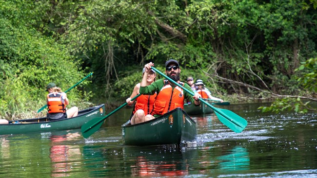 a group of canoeists on the water in green canoes, with 2 person in each canoe