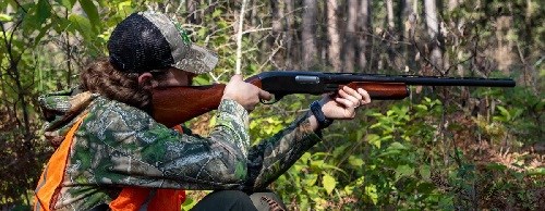 Person in camouflaged shirt, hat, & orange vest kneels in woods with a rifle pointed on his shoulder.