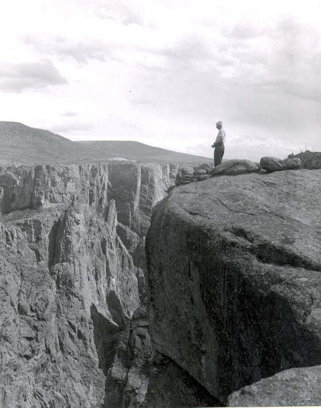 Black and white historic image of a person standing on a canyon rim. Steep cliffs are visible below and beyond the person.