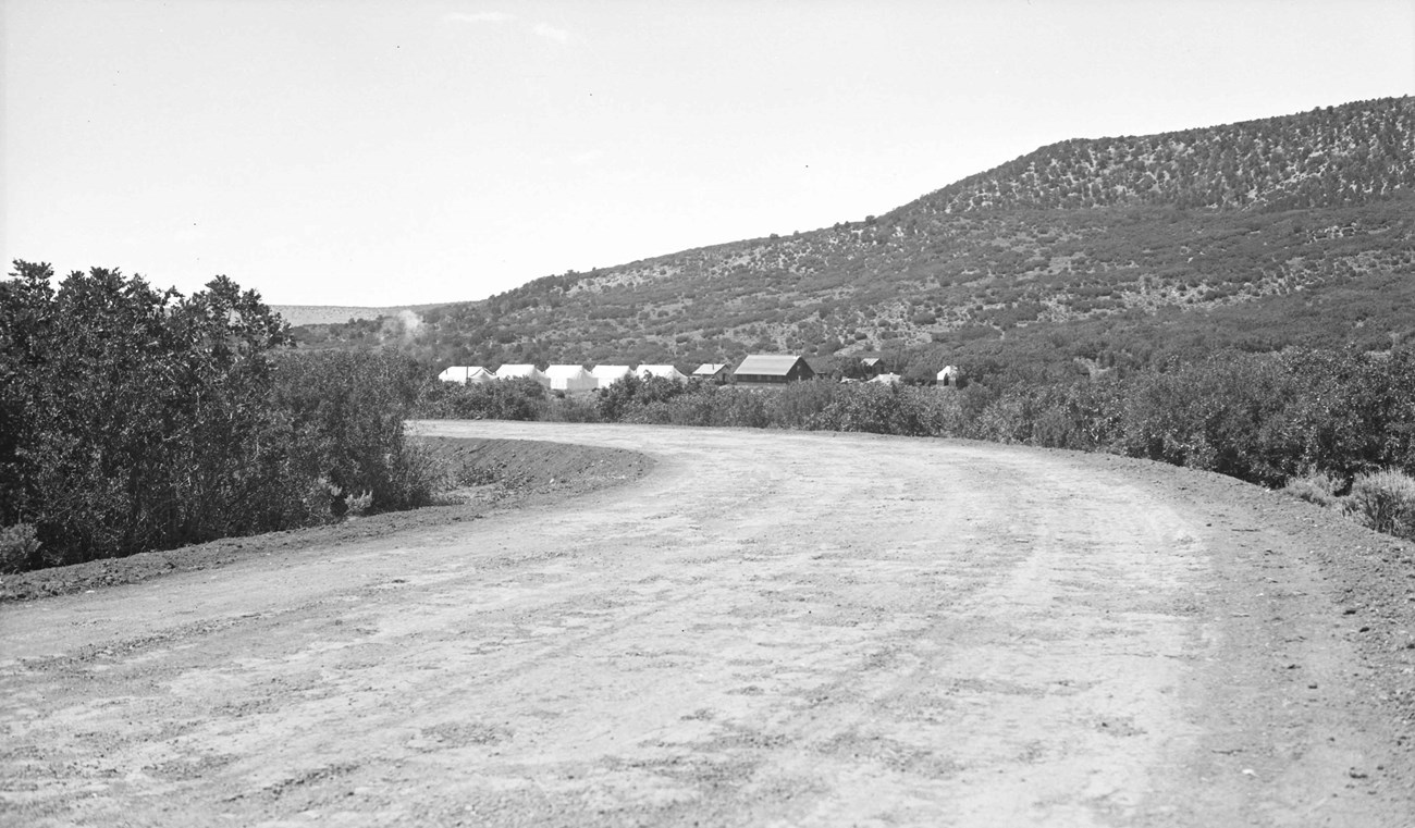Historic image of a road with short vegetation on the roadside. Small simple camp buildings are in the distance. Mesa rises behind the buildings.