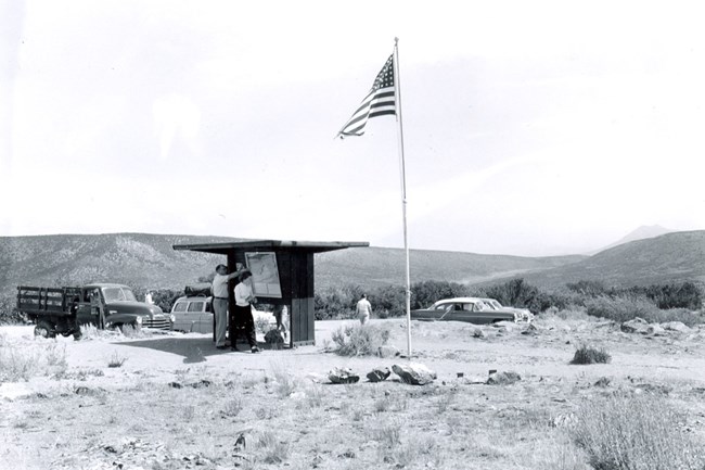 Black and white image showing people standing next to a wooden shelter structure with information panels on it. A flag on a pole is beside it. Cars are parked behind the shelter.