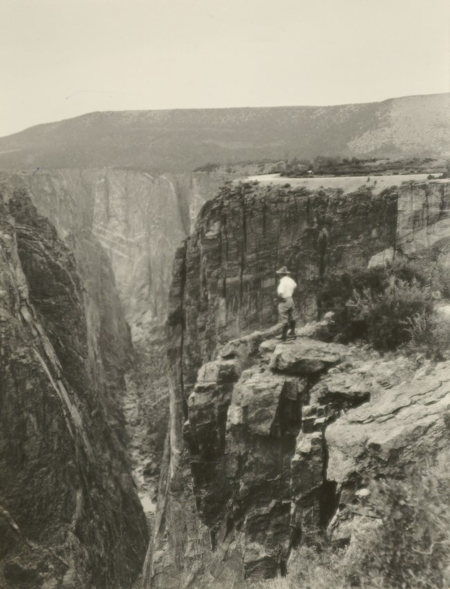Historic black and white image of a person wearing a white shirt and light pants stands by a canyon rim edge. Tall steep canyon walls continue into the distance. A mesa rises above the canyon.