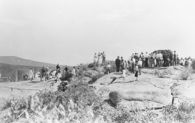 Historic black and white image of a group of people wearing period clothing for the 1930s. A canyon and mesas are beyond the people.