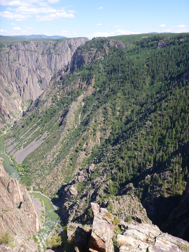 Deep canyon in a "V" shape with more vegetation and conifer trees on the north-facing slopes. A narrow river is visible at the canyon bottom.