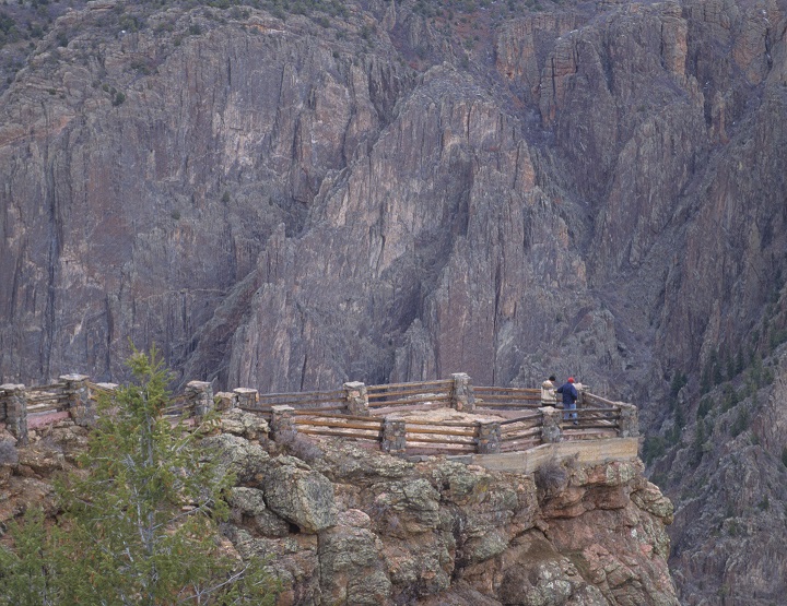 Two visitors stand on edge of deep, dark canyon at a log fence viewpoint Two visitors stand on edge of deep, dark canyon at a log fence viewpoint