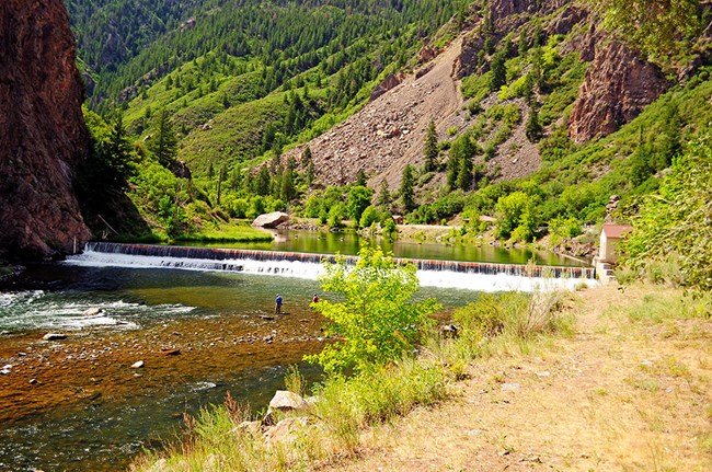 two fishermen wading in shallow river below a small waterfall, gently sloping canyon walls on both sides