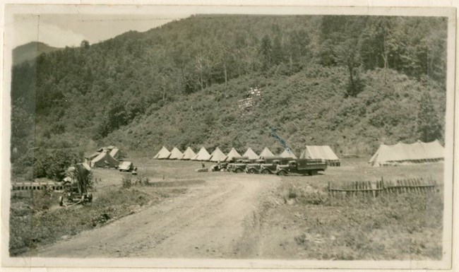 Historic photo of dirt road leading to white canvas tents and buildings. Mountain ridge behind tents.