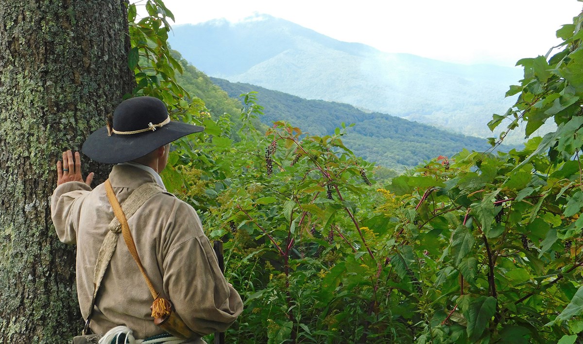 A man in revolutionary era clothing stands next to a tree overlooking mountains and a valleys