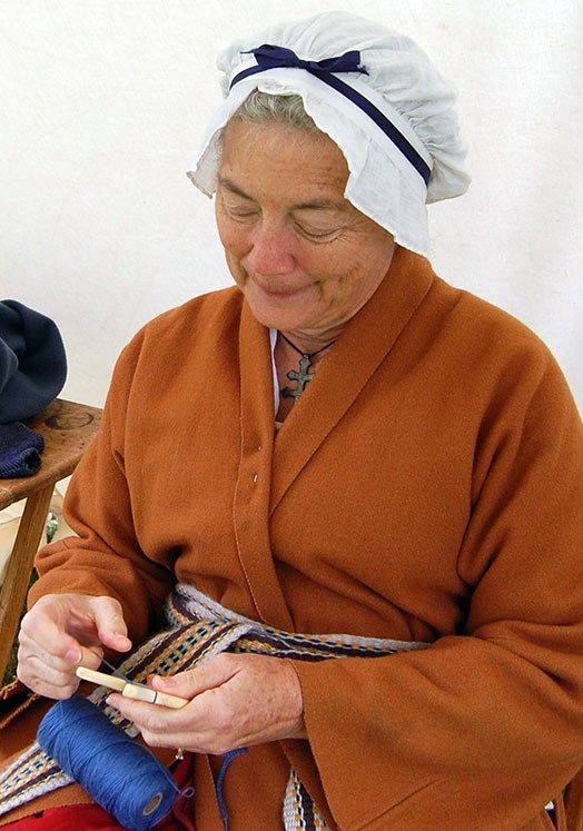 A woman in revolutionary war era clothing sits holding a spool of yarn and a handheld frame for making lucets.