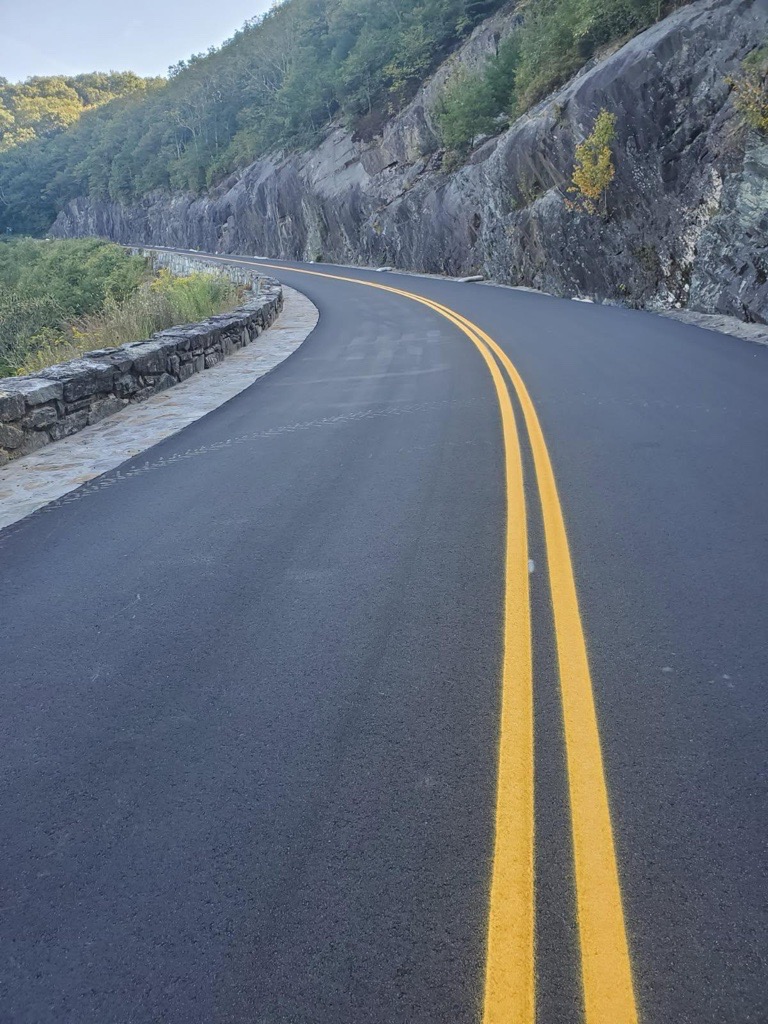 Freshly paved mountain road with double yellow lines curving beside a stone wall and steep, tree-covered rock face