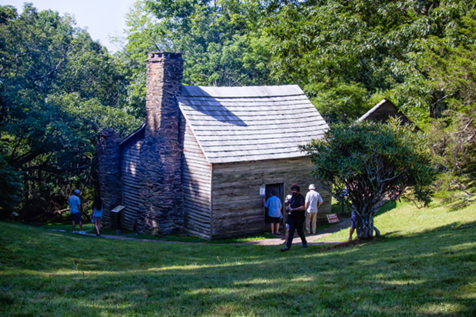 Visitors to the Brinegar cabin observe the displays and architecture of the historic building Visitors gather around an older brick and lumber building in a shady grove