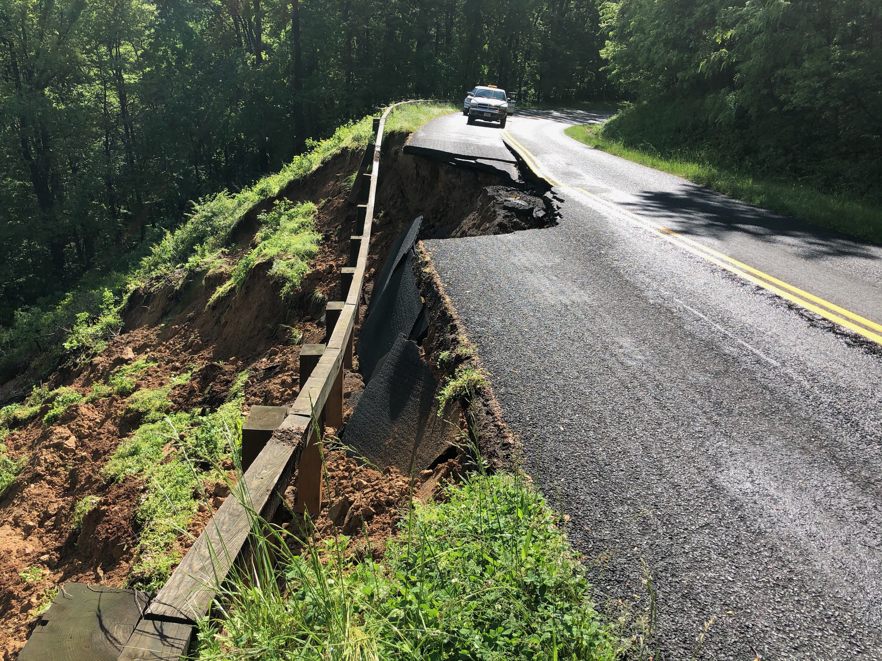 A car parked in front of a road that has sloughed down slope.