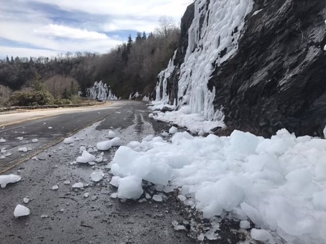 Chunks of ice fall onto the road from a steep cliff on the road shoulder.
