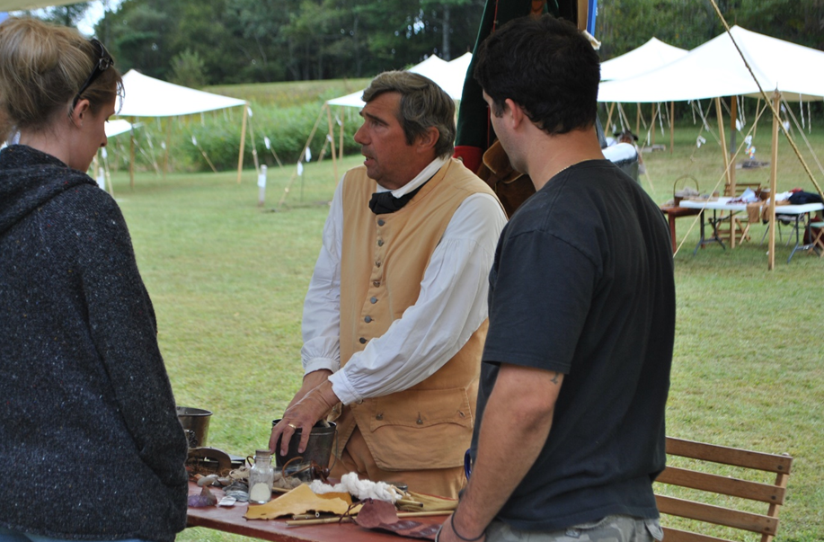 A historic reenactor in colonial garb stands behind a table with antique items while speaking to visitors
