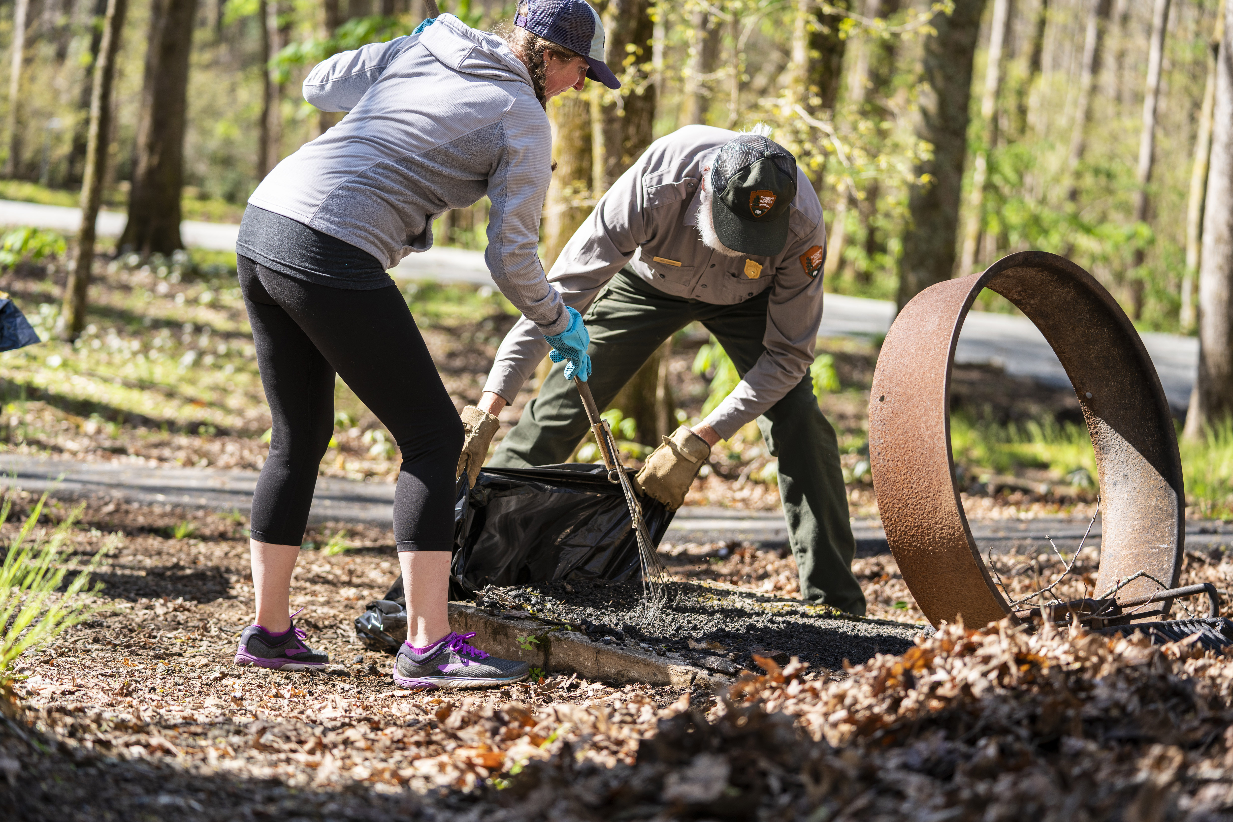 A woman rakes debris into a trash bag held by a park ranger.