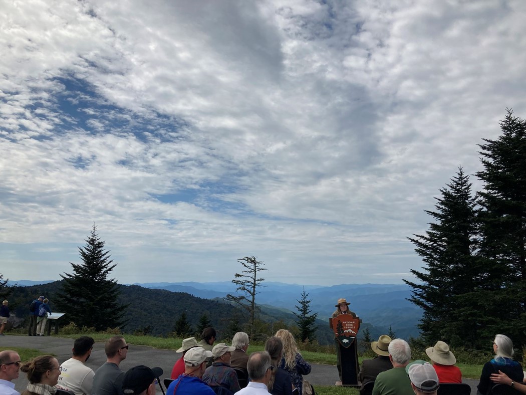 Uniformed National Park Service member stands at a NPS podium in front of a gathered crowd with a sprawling mountain vista in the background