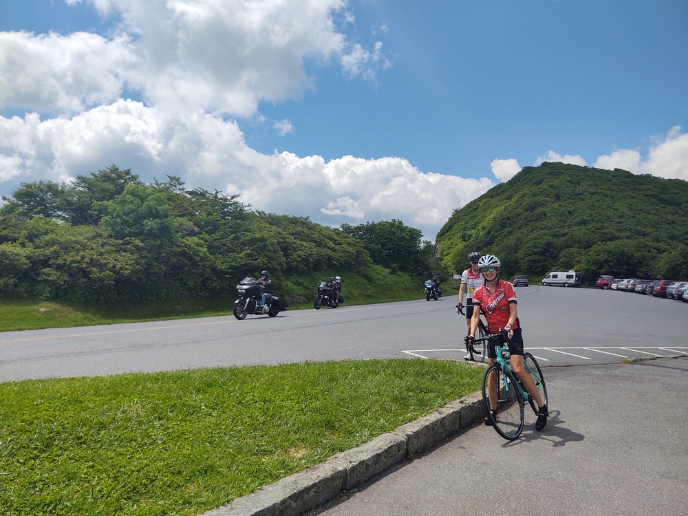 A woman stands near her bicycle next the a curving road.