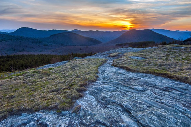 A slick rock hiking trail runs through a grassy alpine meadow with a winter sunset over the mountains on the horizon.