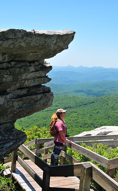 A female hiker with a backpack looks out over the landscape from a wooden hiking boardwalk.